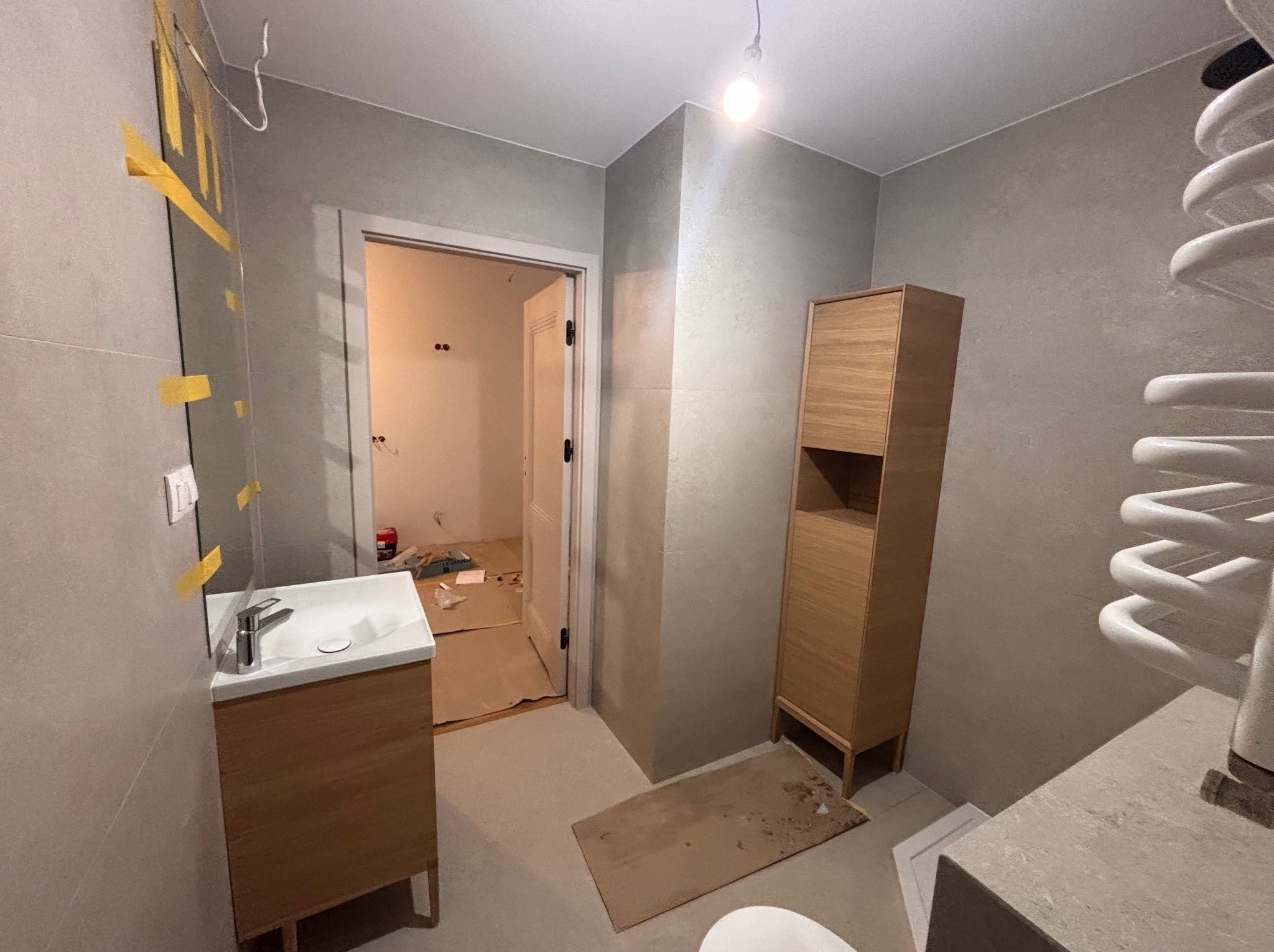 Wnętrze łazienki w trakcie wykańczania: zamontowana szafka z umywalką, grzejnik drabinkowy, szafka stojąca. Widoczne zabezpieczenia na lustrze i kartony na podłodze.
