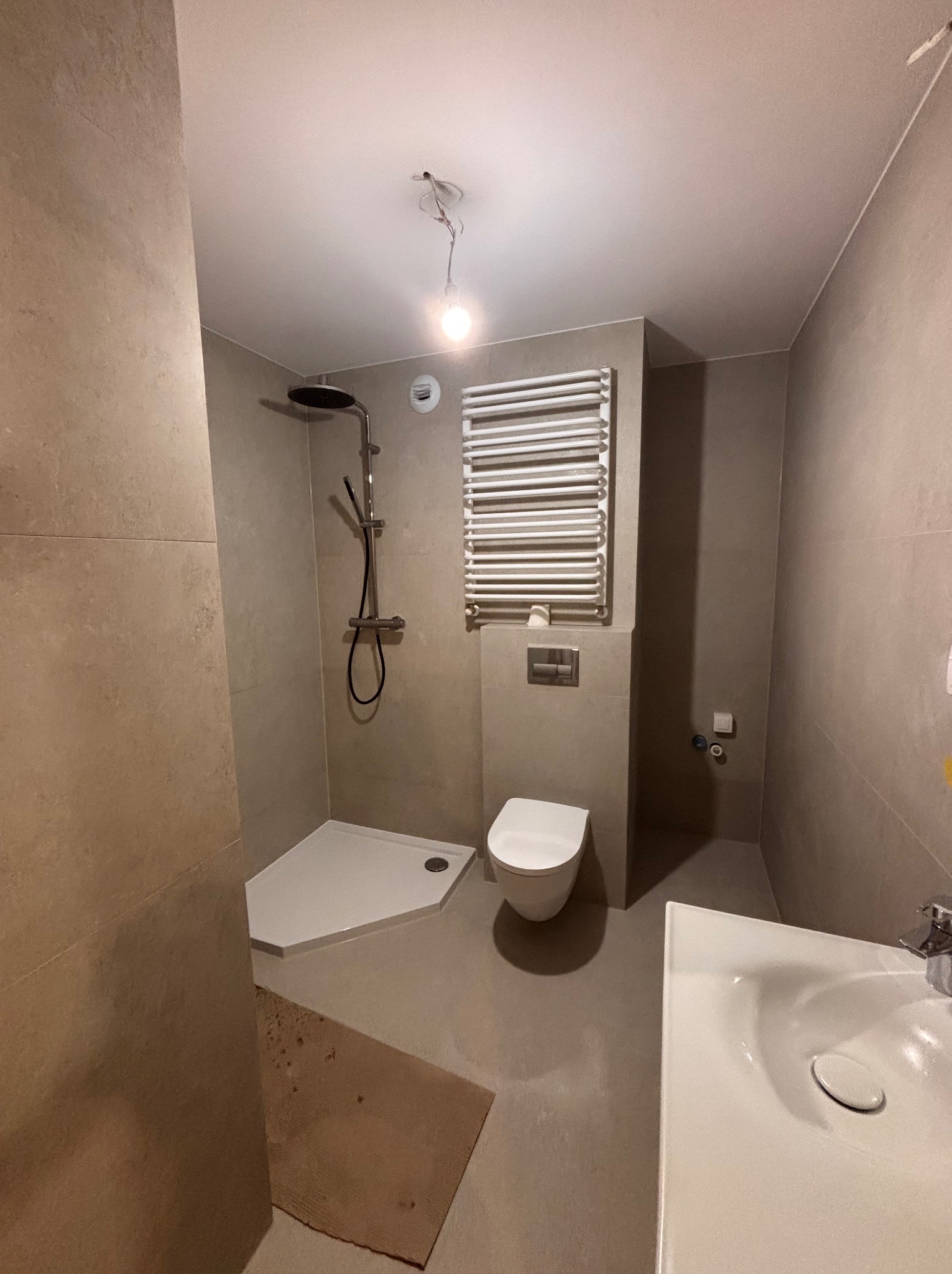 Wykończona łazienka w beżowych tonacjach z kabiną prysznicową, toaletą i grzejnikiem drabinkowym. Widoczne płytki na ścianach i podłodze, minimalistyczny design.