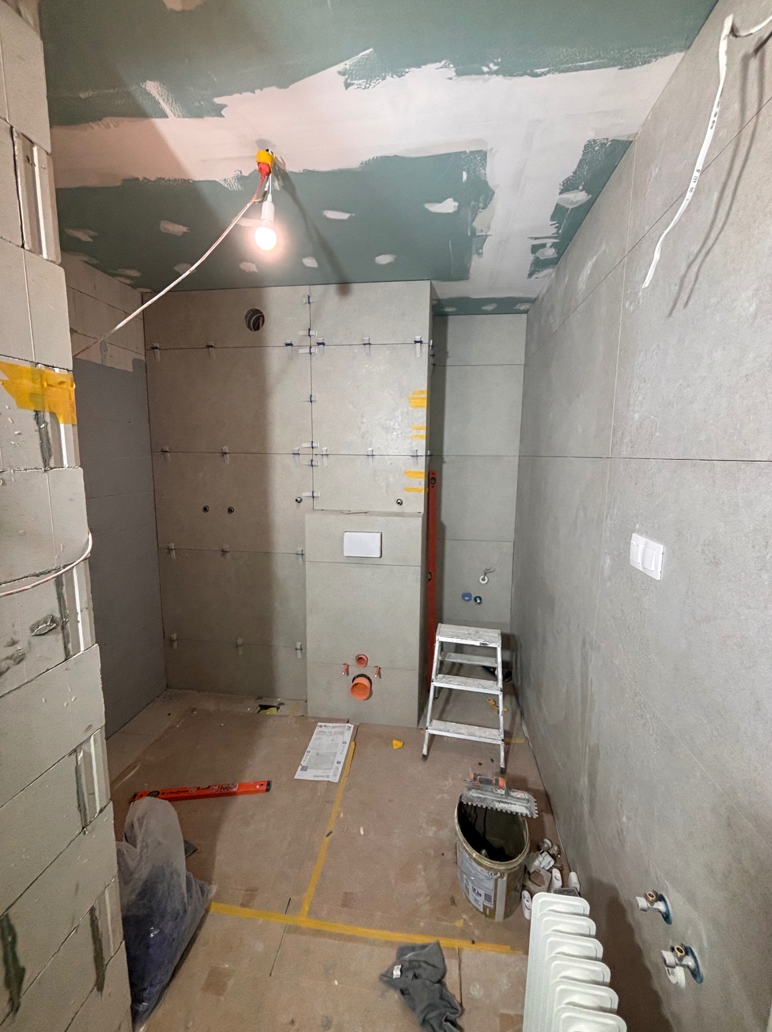Wnętrze łazienki w trakcie remontu: szare płytki na ścianach, widoczne krzyżyki dystansowe, przygotowane podłoże sufitu, drabina, narzędzia i materiały budowlane.