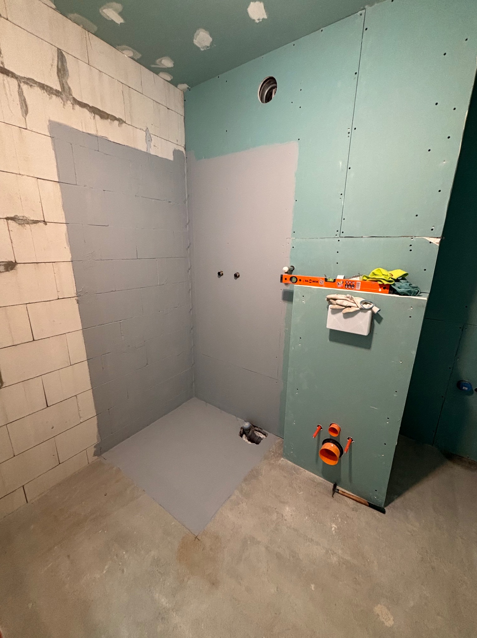 Wnętrze łazienki w trakcie prac: ściany z płyt gips-karton, fragment ściany z bloczków, częściowo pokryte szarą warstwą. Widoczne narzędzia i instalacje.