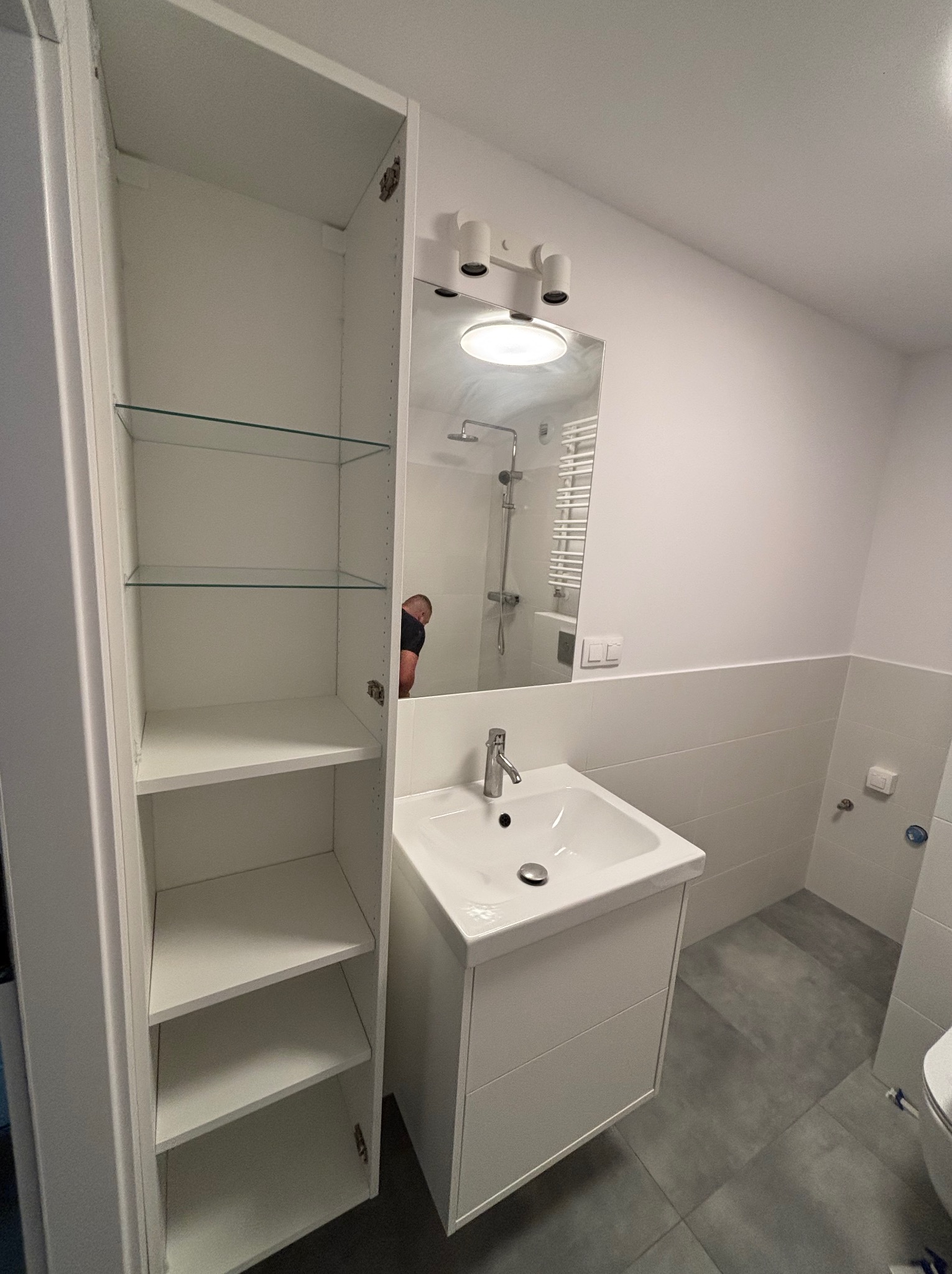 Biała, minimalistyczna łazienka po remoncie: szafka z umywalką, lustro z oświetleniem i wysoka szafka z półkami. Szare płytki podłogowe. Widoczne odbicie osoby w lustrze.