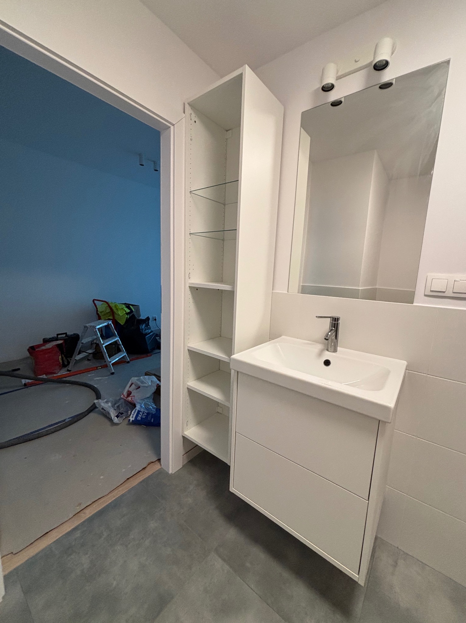 Nowoczesna łazienka w trakcie wykańczania: biała szafka z umywalką, lustro z oświetleniem, regał z półkami. W tle widoczne narzędzia i materiały budowlane.