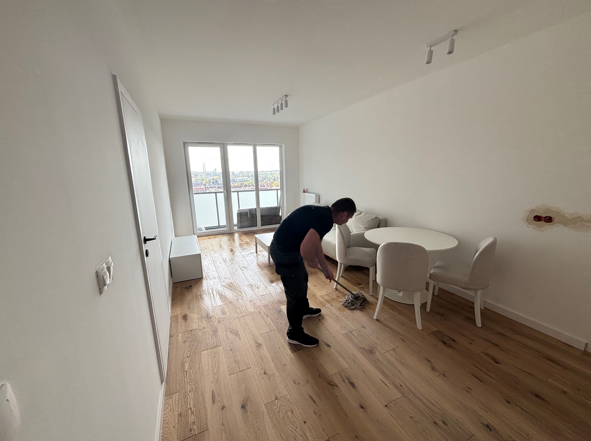 Mężczyzna myje drewnianą podłogę w jasnym, minimalistycznym wnętrzu z balkonem i widokiem na miasto. Widoczne ślady wilgoci po myciu.