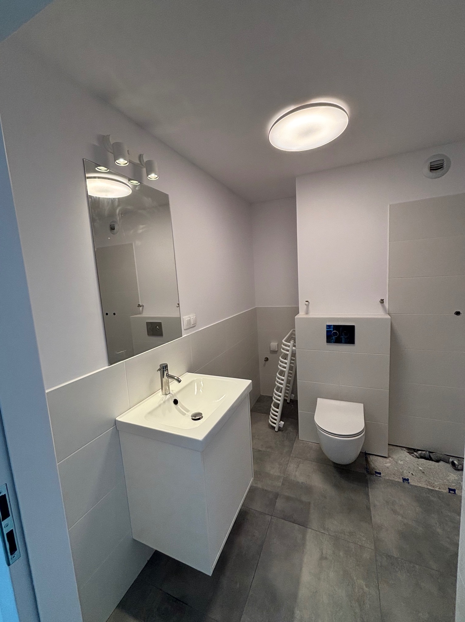 Nowoczesna, biała łazienka po remoncie z umywalką, lustrem i toaletą. Widoczne szare płytki podłogowe i białe ściany. Minimalistyczny design z oświetleniem sufitowym.