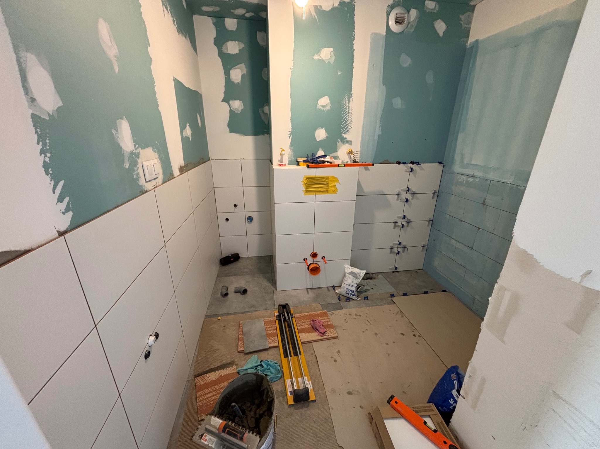 Wnętrze łazienki w trakcie prac. Częściowo położone białe płytki na ścianach, widoczne narzędzia glazurnicze, ślady po szpachlowaniu na ścianach.