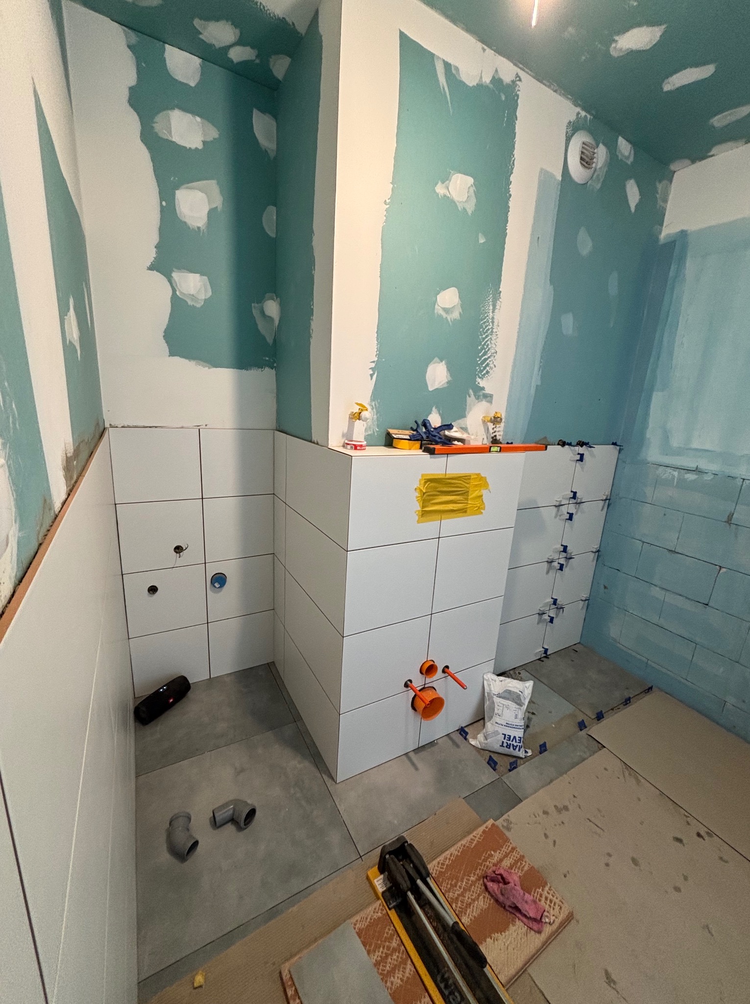 Wnętrze łazienki w trakcie prac wykończeniowych: białe płytki na ścianach, szpachlowane ściany w odcieniach błękitu, narzędzia i materiały budowlane widoczne na podłodze.