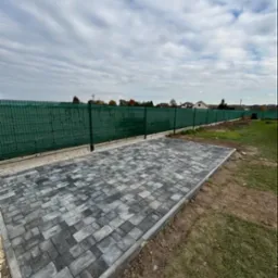 Ułożona nawierzchnia z kostki brukowej w odcieniach szarości, zakończona obrzeżem, widok z góry na tle zielonego ogrodzenia i pochmurnego nieba.