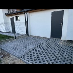 Alan Głuchowski - Szare ażurowe płyty betonowe tworzące nawierzchnię obok budynku z szarą stolarką drzwiową i okienną, widoczne elementy obrzeża i fragment trawnika.