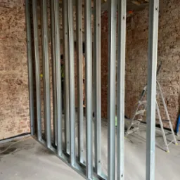 Stelaż z profili stalowych do zabudowy ścianki działowej z płyt gipsowo-kartonowych na tle ściany z czerwonej cegły, widoczna drabina aluminiowa i niebieska rura.