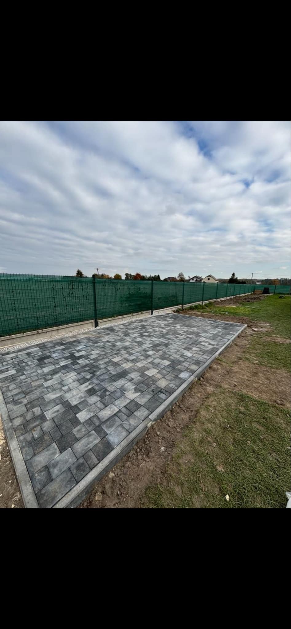 Ułożona nawierzchnia z kostki brukowej w odcieniach szarości, zakończona obrzeżem, widok z góry na tle zielonego ogrodzenia i pochmurnego nieba.