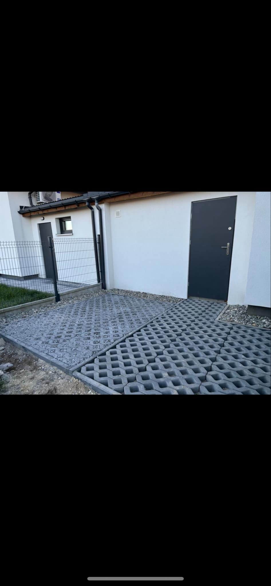 Szare ażurowe płyty betonowe tworzące nawierzchnię obok budynku z szarą stolarką drzwiową i okienną, widoczne elementy obrzeża i fragment trawnika.