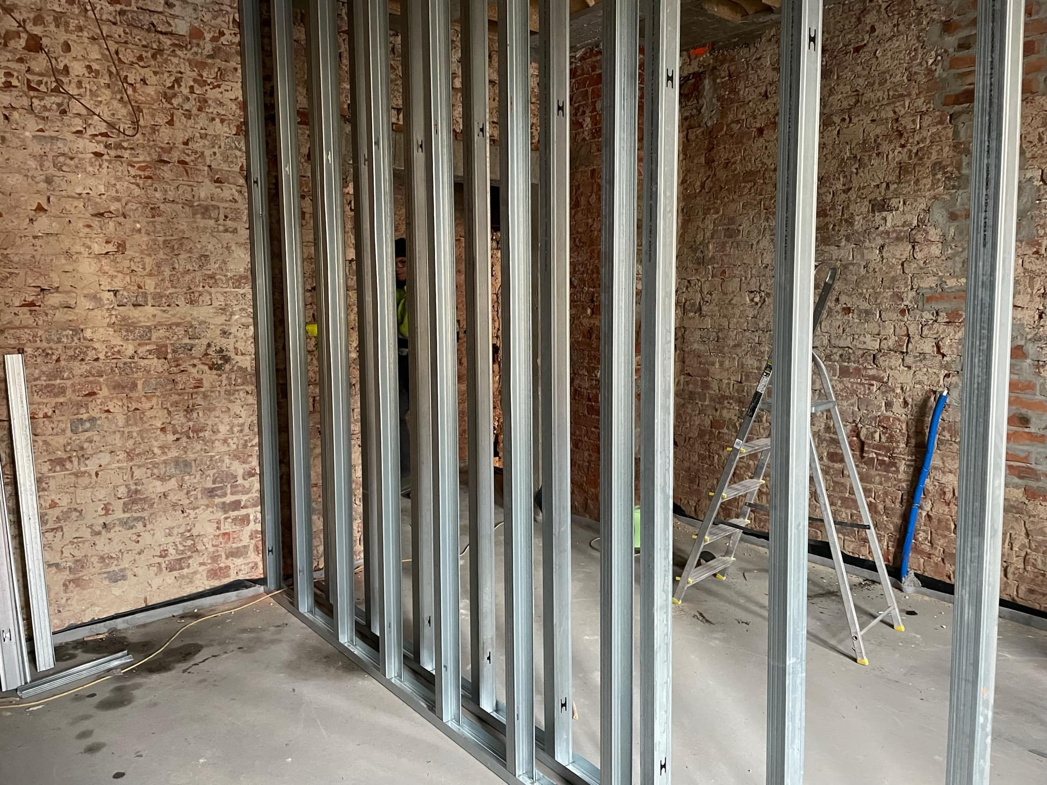 Stelaż z profili stalowych do zabudowy ścianki działowej z płyt gipsowo-kartonowych na tle ściany z czerwonej cegły, widoczna drabina aluminiowa i niebieska rura.