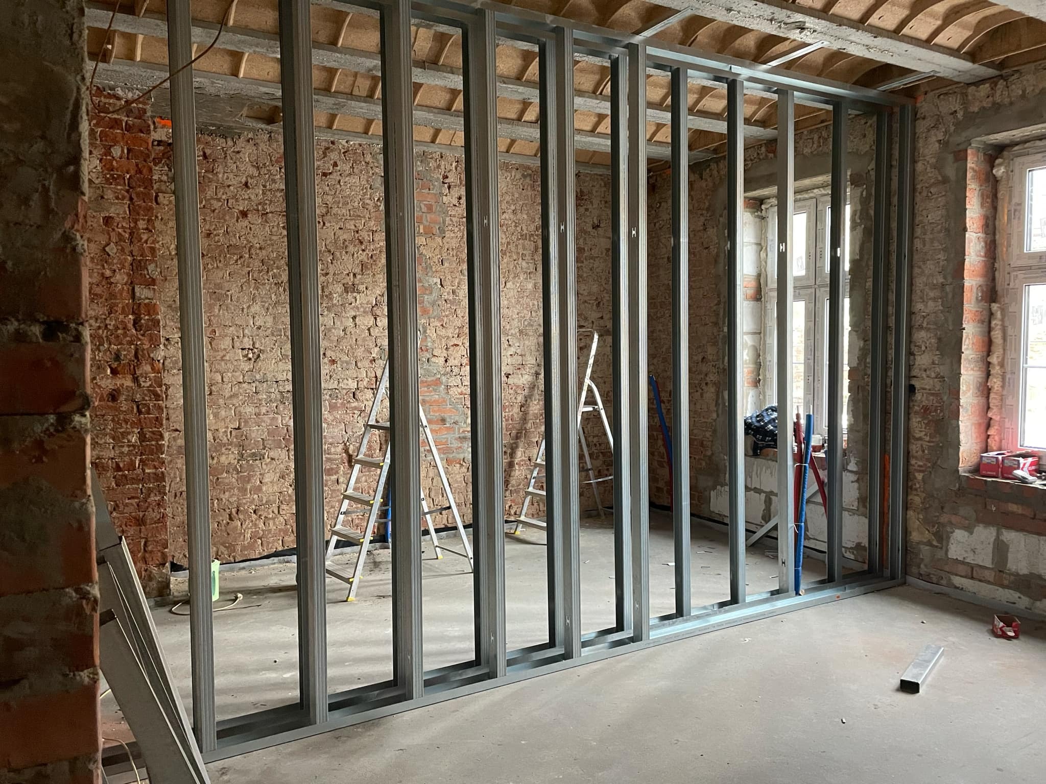 Stelaż z profili stalowych do ścianki działowej, widoczny na tle surowych, ceglanych ścian w trakcie remontu. Dwie drabiny aluminiowe w tle. Instalacje wodne i elektryczne przygotowane do montażu.
