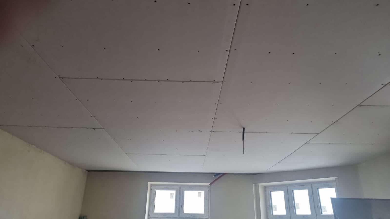 Montaż sufitu podwieszanego z płyt karton gips z widocznymi łączeniami i wkrętami, instalacja elektryczna wystająca z sufitu, okna PCV w tle.