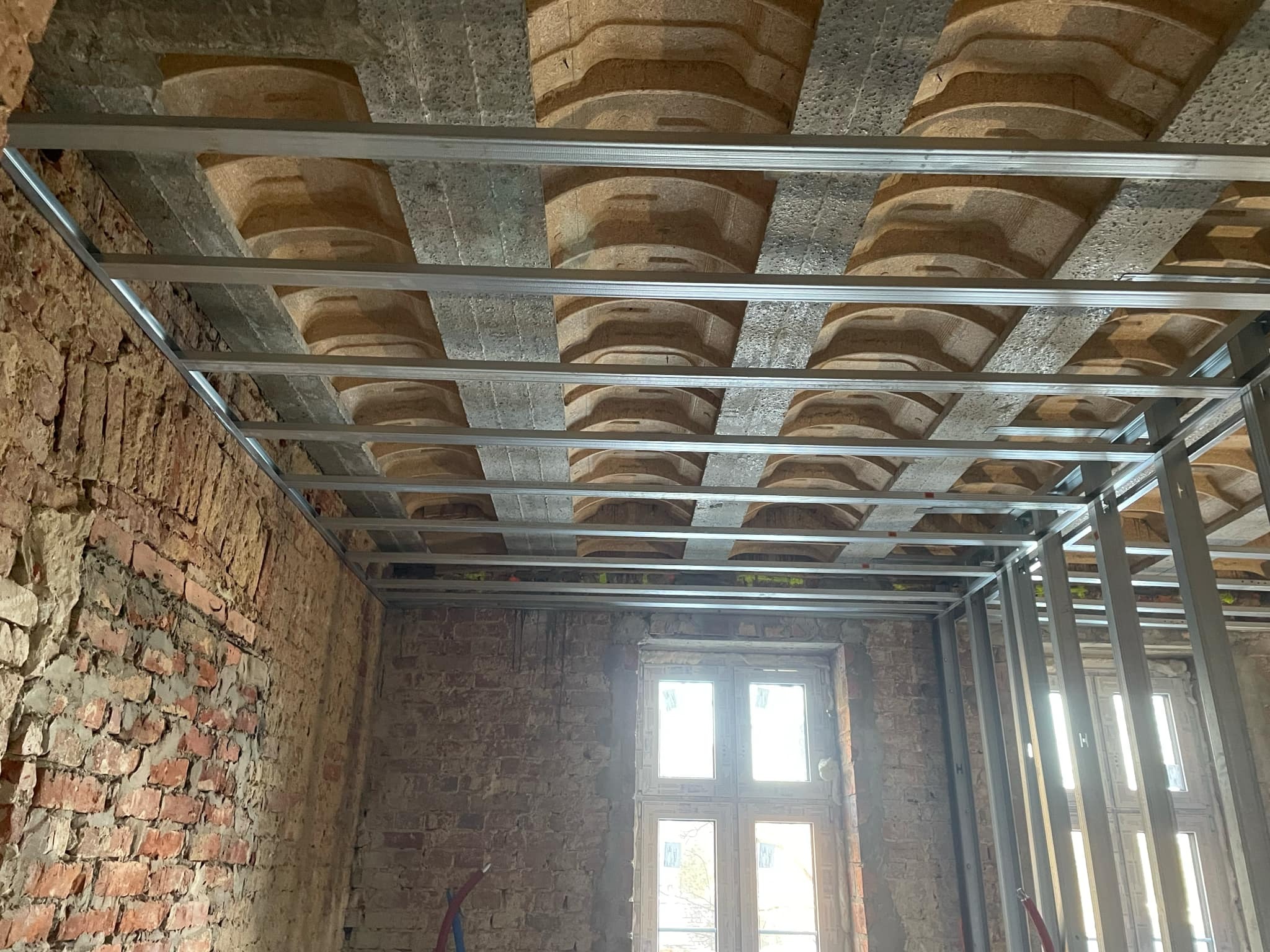 Metalowa konstrukcja sufitowa w trakcie montażu, widoczne stare ceglane ściany i fragmenty stropu z reliefami, okno w tle.