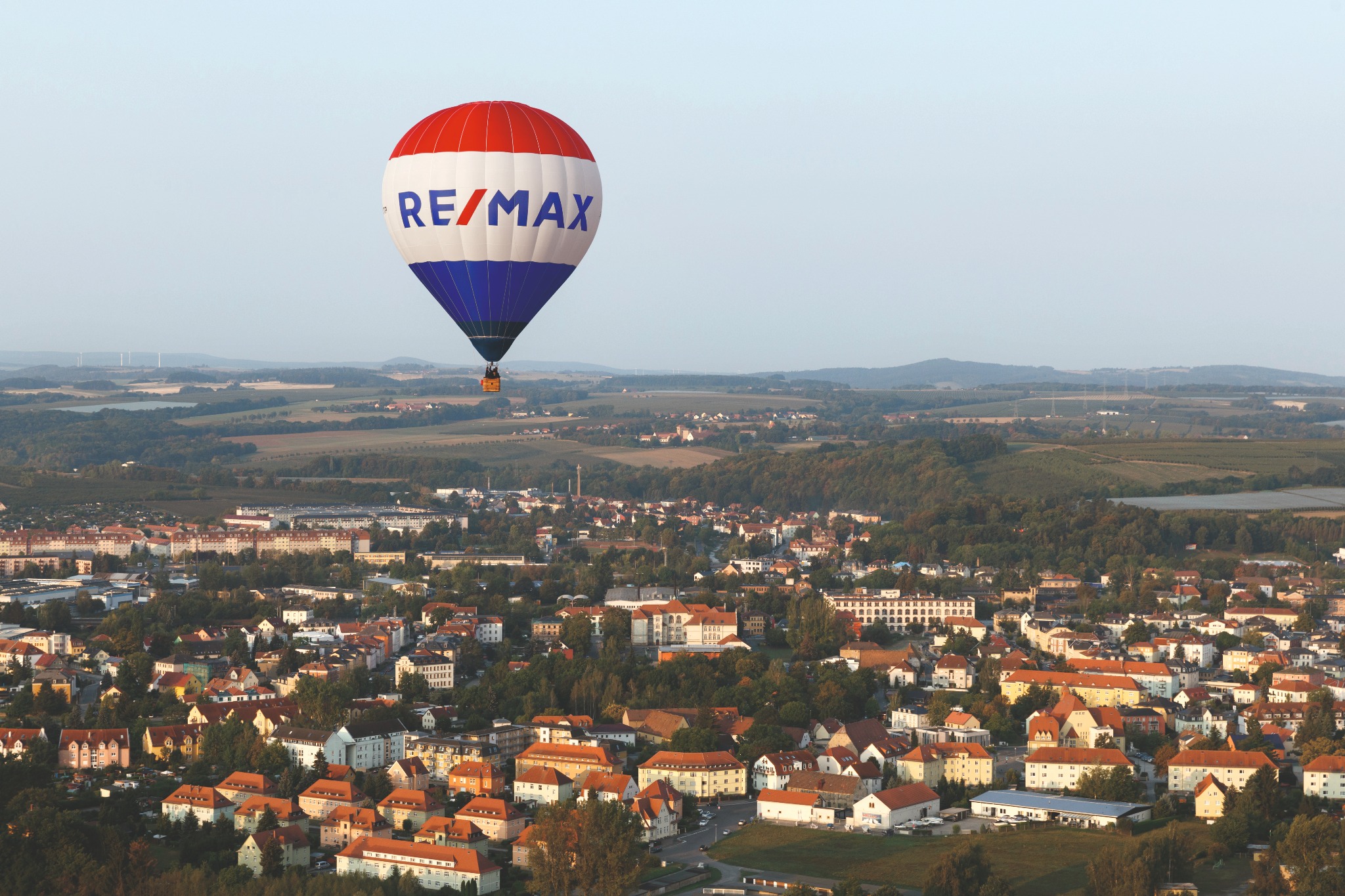 Balon z logo RE/MAX nad miastem, widok z góry na zabudowę mieszkalną i tereny zielone w oddali.