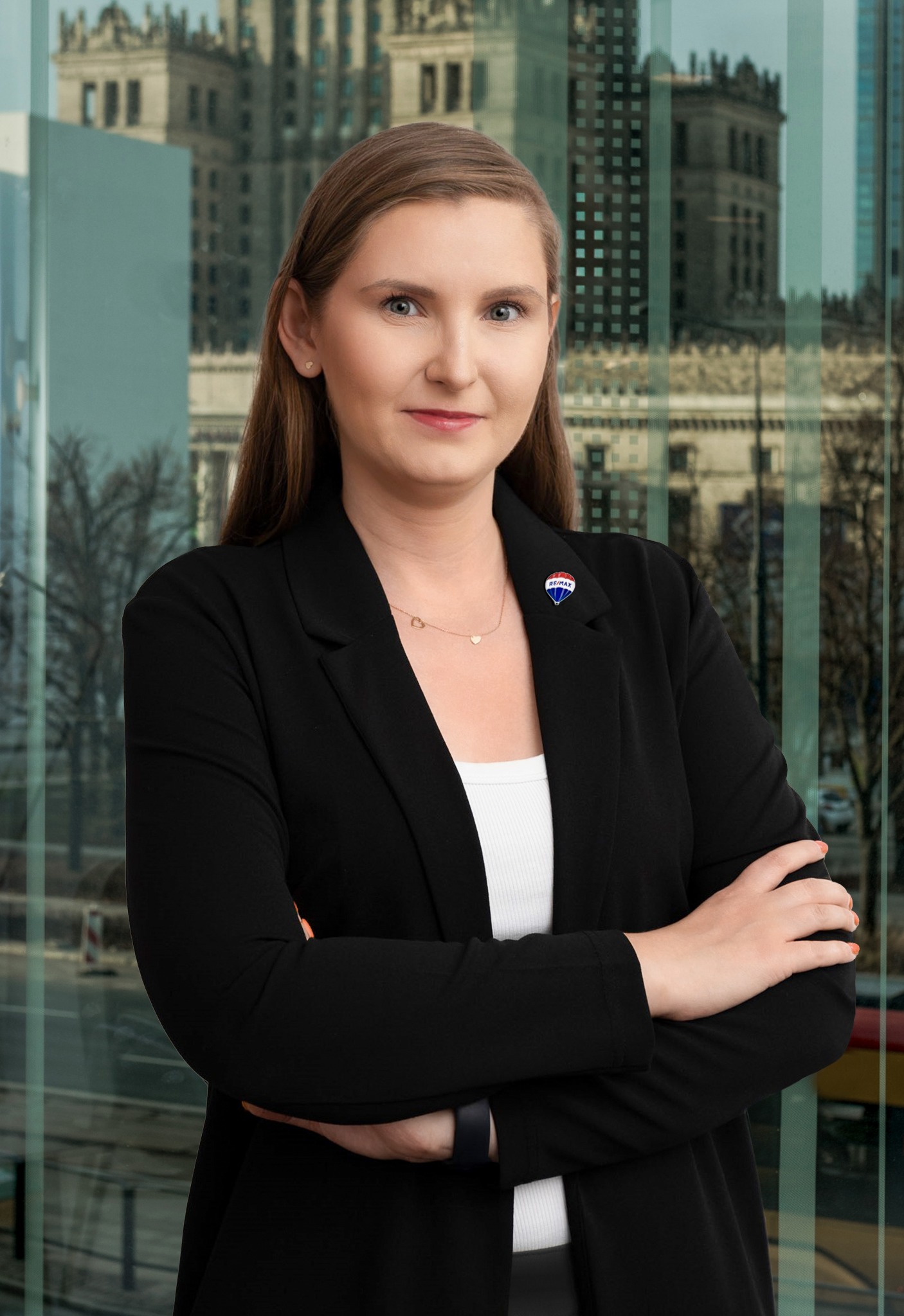 Profesjonalny portret kobiety w czarnym żakiecie z przypinką RE/MAX, na tle okna z widokiem na Pałac Kultury w Warszawie.