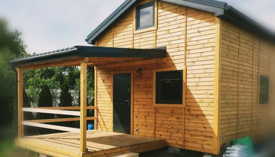 Gotowy domek letniskowy z drewna, z zadaszonym tarasem i ciemną stolarką okienną, widoczne worki z materiałami budowlanymi obok.