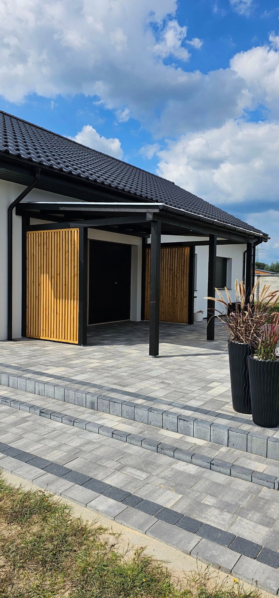 Nowoczesny dom z czarną dachówką i drewnianymi panelami osłonowymi, widok na szary podjazd z kostki brukowej i ozdobne donice z trawami.