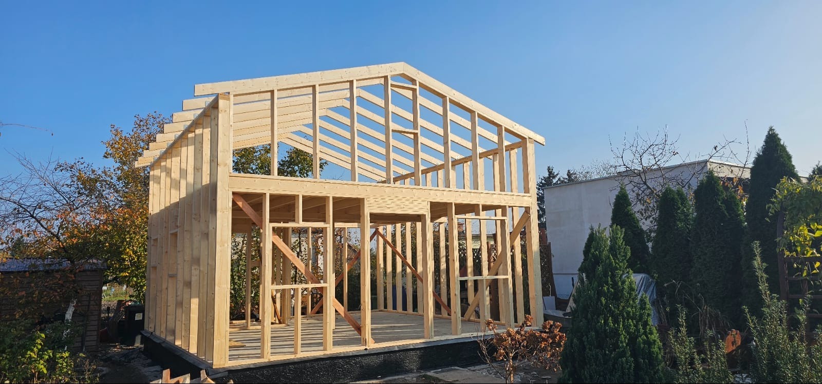 Surowy szkielet drewnianego domu w trakcie budowy, widoczne elementy konstrukcyjne dachu i ścian, błękitne niebo w tle.