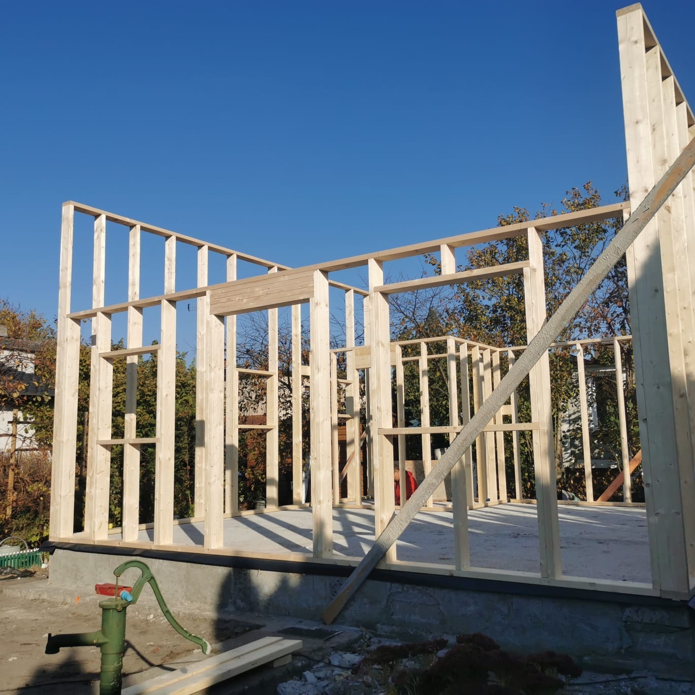 Wznoszenie konstrukcji drewnianej domu z widoczną ramą ścian, słupami i belkami na fundamencie, z zieloną pompą ręczną do wody w tle, pod błękitnym niebem.