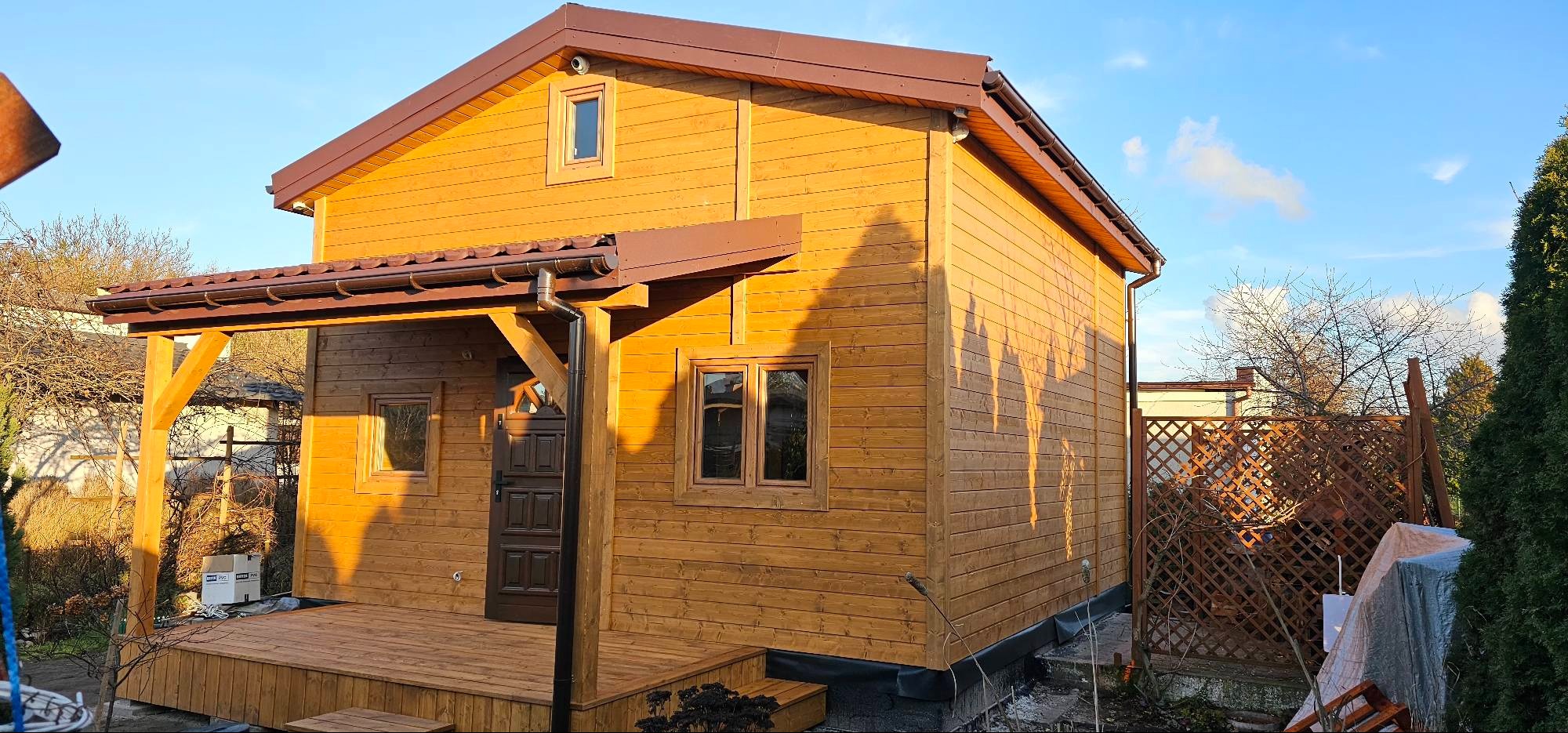 Drewniany dom szkieletowy z brązowym dachem i zadaszonym tarasem, widok z zewnątrz w słoneczny dzień.