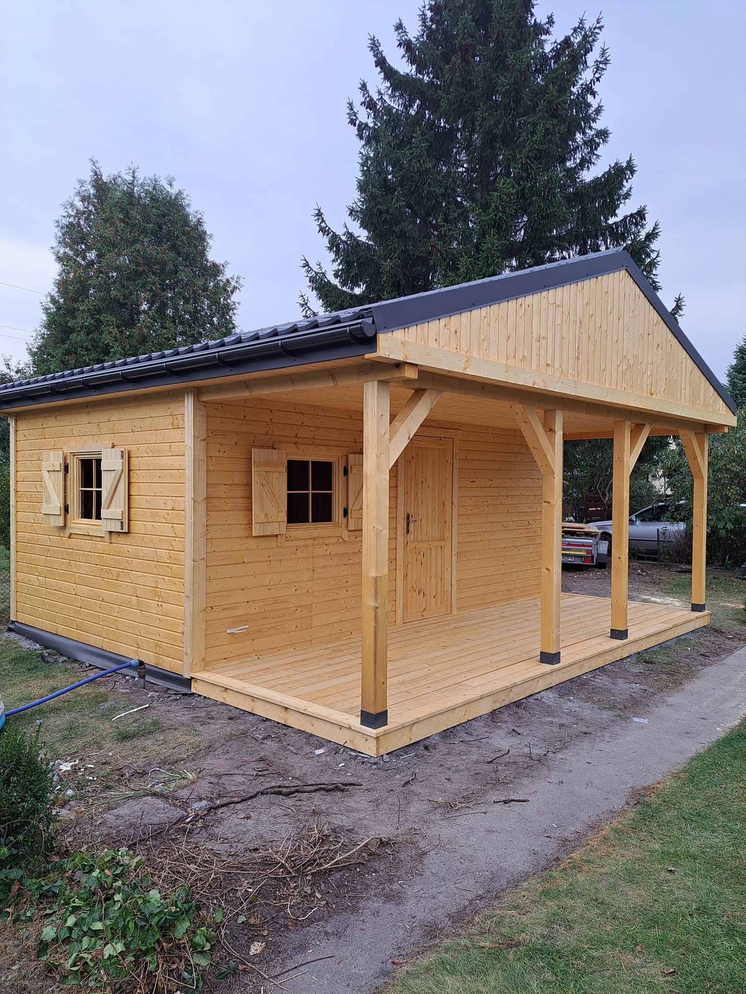 Drewniany domek letniskowy z zadaszonym tarasem, okiennice otwarte, widok z boku na tle drzew i pochmurnego nieba.