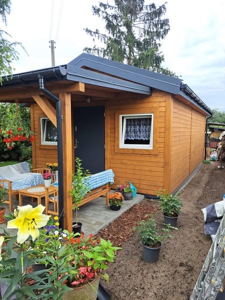 Drewniany domek letniskowy z szarą stolarką i zadaszeniem w Solcu Kujawskim, otoczony rabatami kwiatowymi i meblami ogrodowymi.