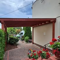 Drewniana wiata garażowa przy domu z jasną elewacją, widok na podjazd z kostki brukowej i rośliny ozdobne, w tle zaparkowany bus.