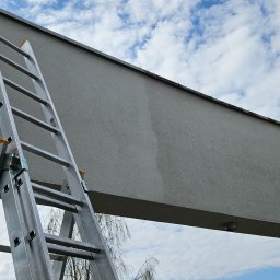 Myjnia Zielona ŁUKASZ KISIELEWSKI - Aluminiowa drabina przystawiona do szarej elewacji budynku na tle błękitnego nieba z obłokami. Widoczne ślady po czyszczeniu na fasadzie.