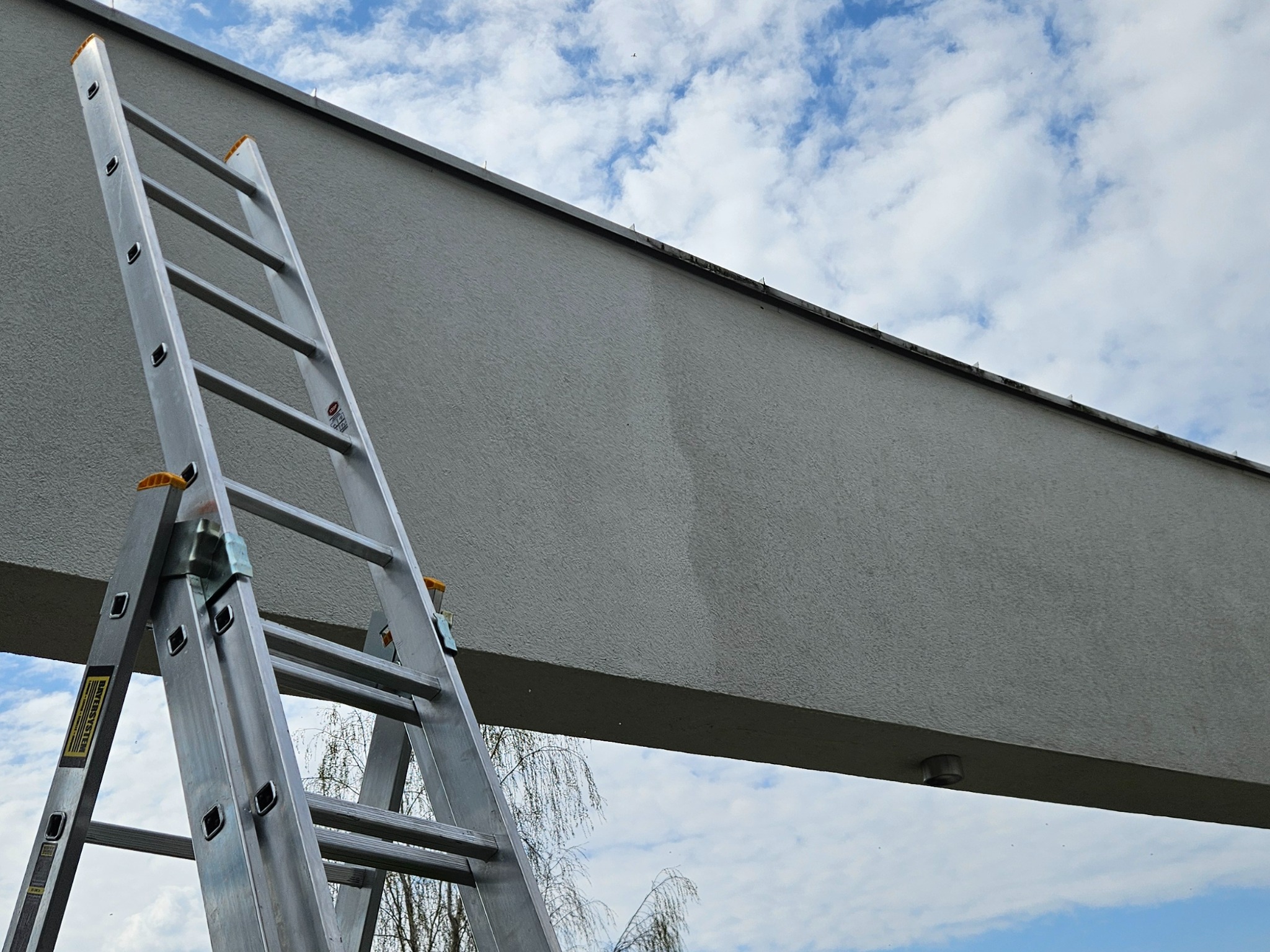 Aluminiowa drabina przystawiona do szarej elewacji budynku na tle błękitnego nieba z obłokami. Widoczne ślady po czyszczeniu na fasadzie.
