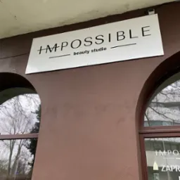 Szyld reklamowy 'IMPOSSIBLE beauty studio' na brązowej ścianie budynku, widoczny z poziomu ulicy. W tle okna z widokiem na drzewa.