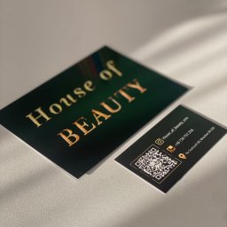 Magnetic design - Dwie czarno-złote wizytówki 'House of Beauty' z kodem QR, leżące na teksturowanym podłożu w naturalnym świetle, ukazujące detale projektu i jakość druku.