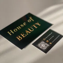 Dwie czarno-złote wizytówki 'House of Beauty' z kodem QR, leżące na teksturowanym podłożu w naturalnym świetle, ukazujące detale projektu i jakość druku.