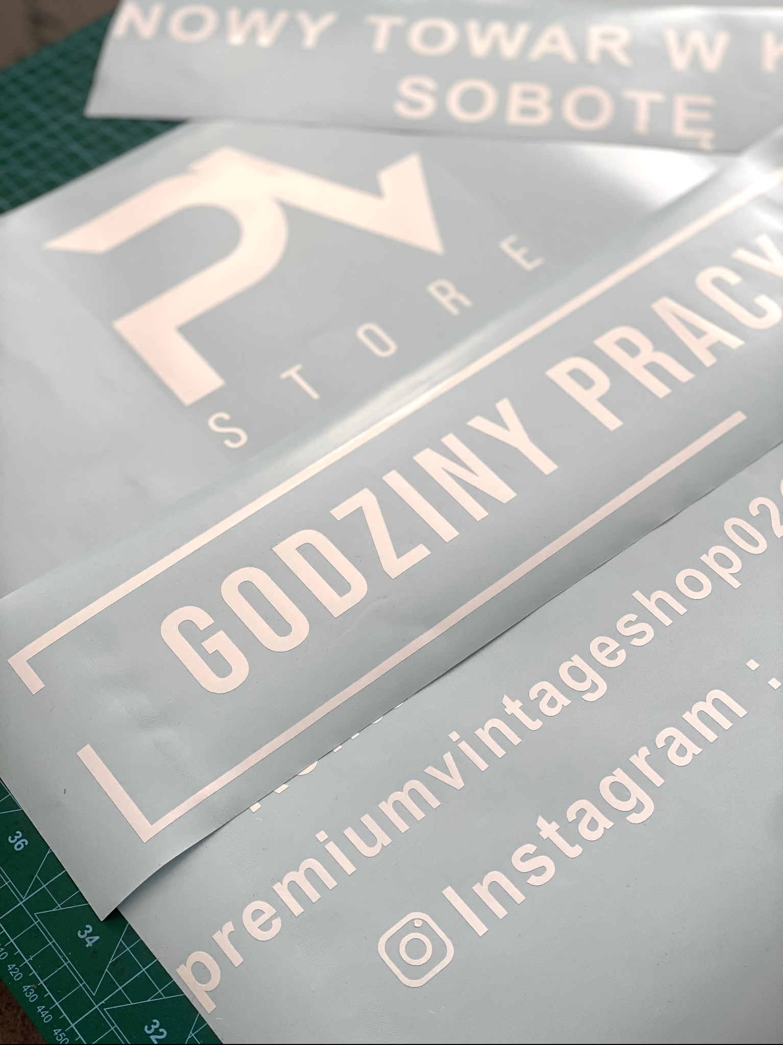 Białe, wycięte napisy na niebieskiej folii, układające się w hasła reklamowe i dane kontaktowe, leżące na zielonej macie. Widoczne logo 'PV STORE' i adres Instagram.