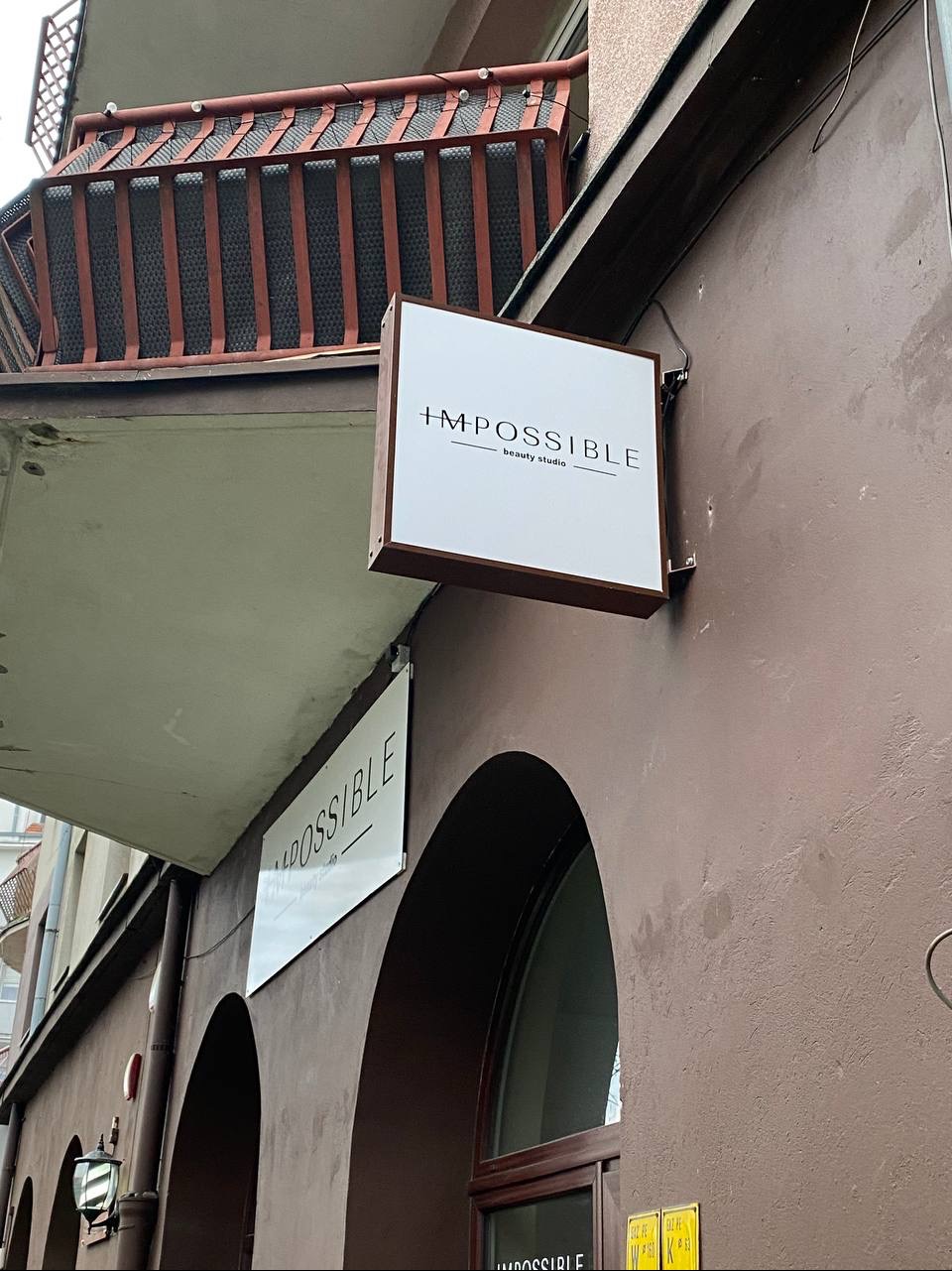 Kwadratowy szyld 'Impossible beauty studio' na brązowej ścianie budynku z balkonem. Ujęcie z dołu, widoczne łuki okien i inne szyldy.