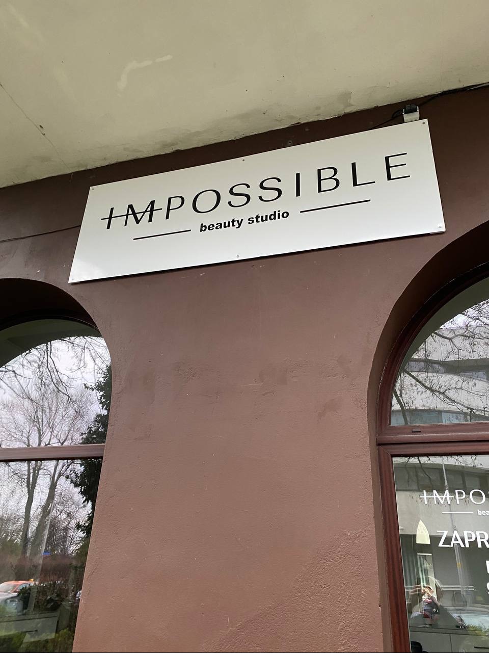 Szyld reklamowy 'IMPOSSIBLE beauty studio' na brązowej ścianie budynku, widoczny z poziomu ulicy. W tle okna z widokiem na drzewa.