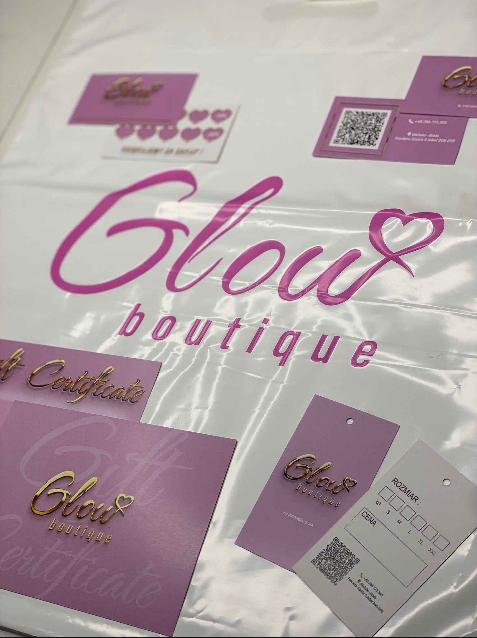 Torba z nadrukiem 'Glow boutique' w odcieniach różu i złota. Widoczne wizytówki, certyfikaty podarunkowe i metki produktowe z logo firmy. Elementy identyfikacji wizualnej.