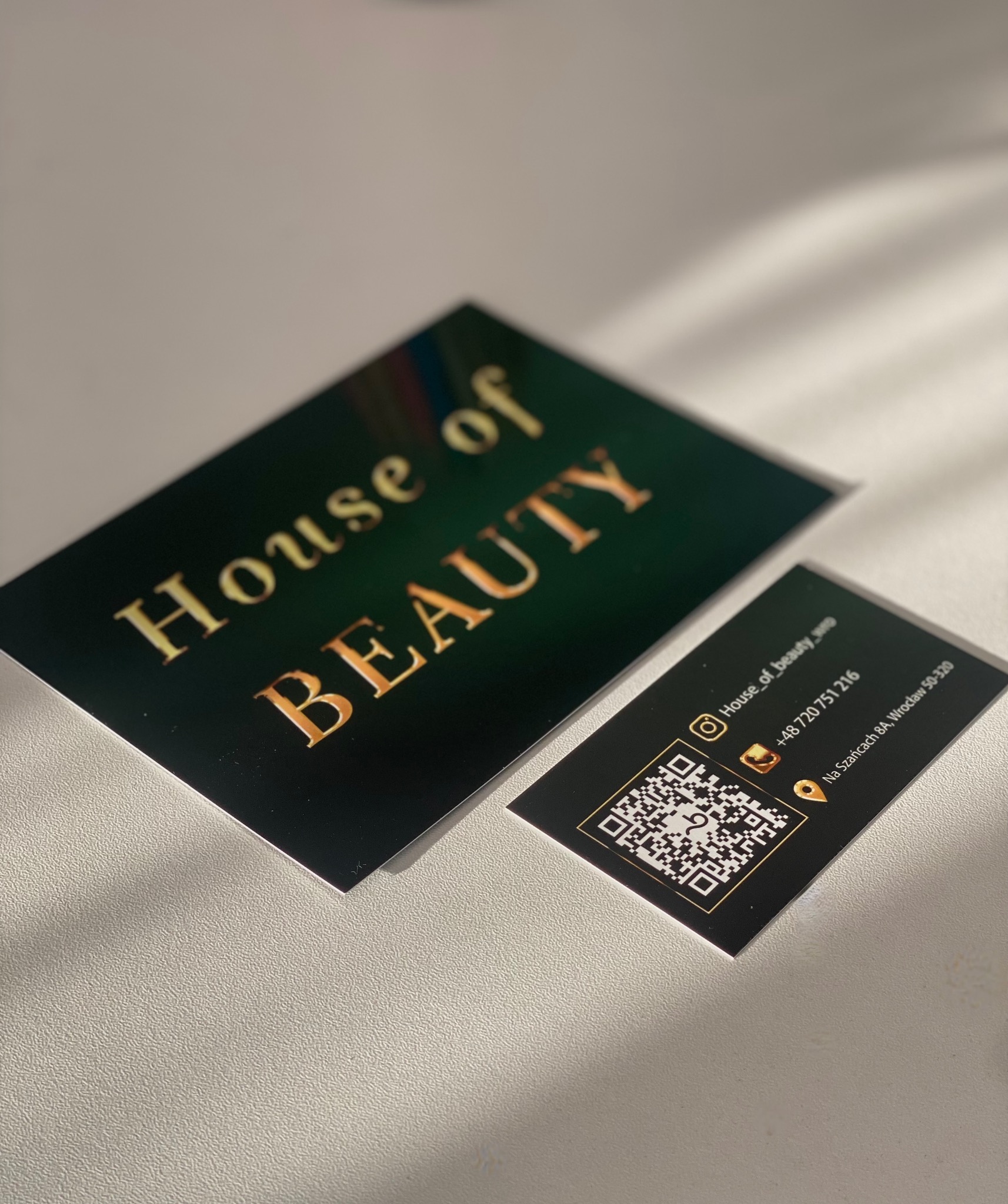 Dwie czarno-złote wizytówki 'House of Beauty' z kodem QR, leżące na teksturowanym podłożu w naturalnym świetle, ukazujące detale projektu i jakość druku.