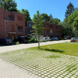 Widok na nowoczesne budynki mieszkalne z czerwonej cegły z ażurowymi elementami, z parkingiem wyłożonym ażurową kostką brukową, przez którą przebija zieleń trawy.