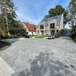 Wrona Investment - Nowoczesny podjazd z szarej kostki brukowej prowadzący do szeregowca z garażem, widoczne drzewa i trawnik.