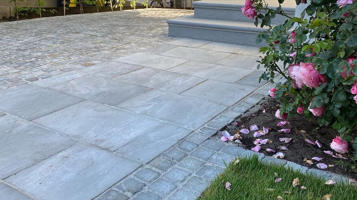 Ułożona nawierzchnia z płyt i kostki brukowej w odcieniach szarości, z elementami zieleni i różowych kwiatów, prowadząca do schodów wejściowych.