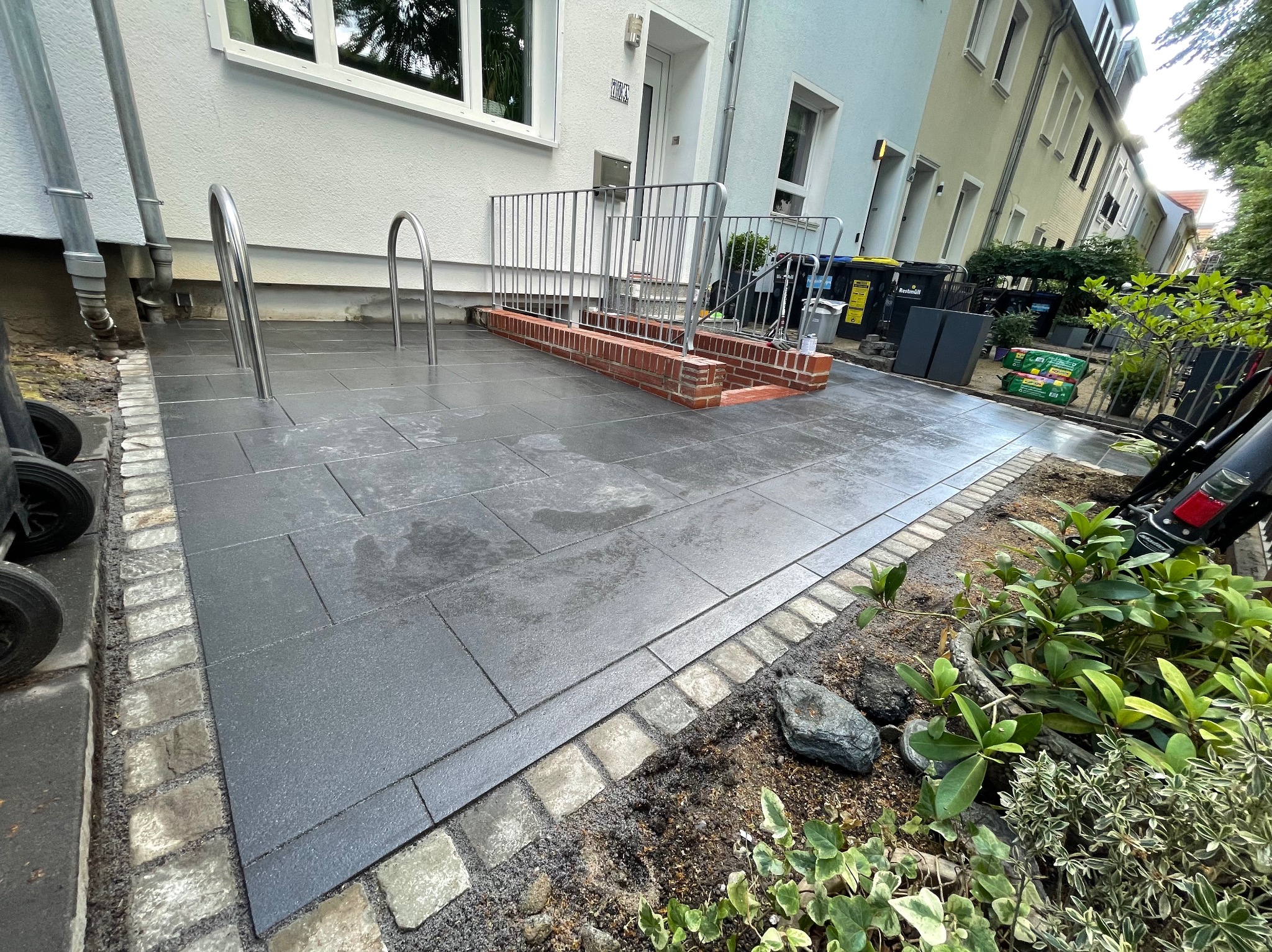 Nowo ułożony, mokry chodnik z szarych płyt, obrzeża z jasnej kostki, metalowe poręcze przy wejściu do budynku, widoczne rośliny wzdłuż chodnika.