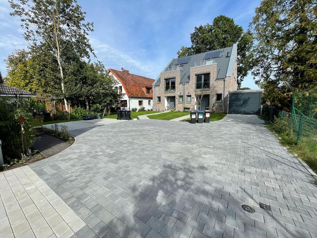 Nowoczesny podjazd z szarej kostki brukowej prowadzący do szeregowca z garażem, widoczne drzewa i trawnik.