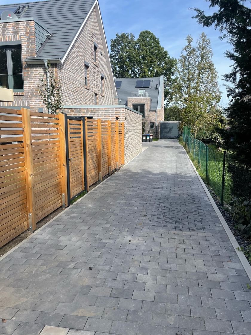 Ułożona szara kostka brukowa na podjeździe, z drewnianym ogrodzeniem po lewej stronie i zielonym ogrodzeniem z siatki po prawej, prowadząca do garażu między dwoma budynkami z cegły.