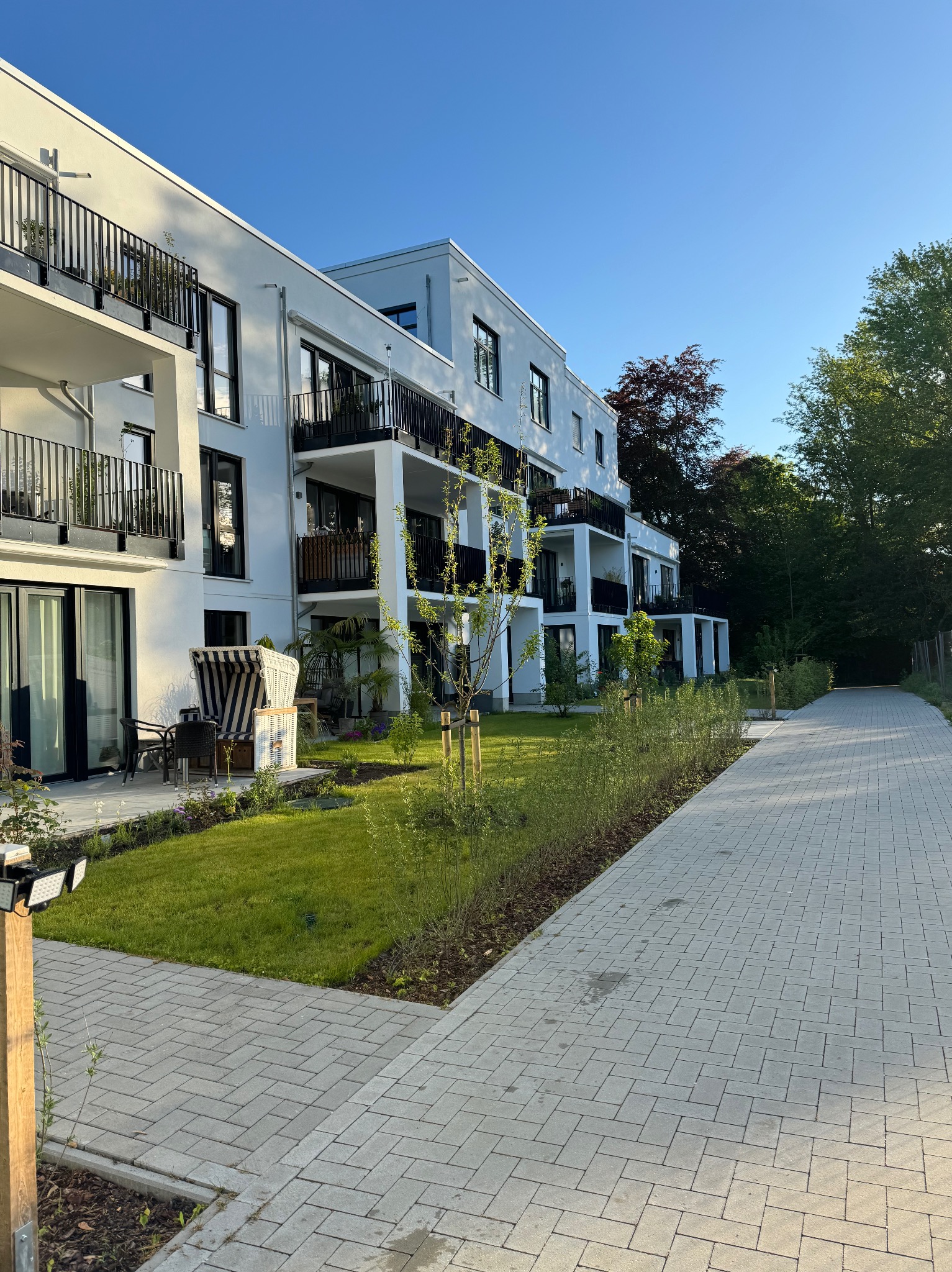 Nowoczesny budynek apartamentowy z balkonami i tarasami, widoczny z poziomu chodnika wyłożonego kostką brukową, z elementami zagospodarowania terenu zielonego i rattanowym koszem plażowym.