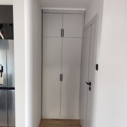 Michał Przybyszewski - Wąski korytarz z szafą wnękową do sufitu w odcieniach szarości i drewnianą podłogą, widoczna częściowo lodówka ze stali nierdzewnej.