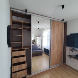 Michał Przybyszewski - Duża szafa przesuwna z lustrzanymi drzwiami i drewnianymi panelami, z otwartą częścią z półkami i szufladami, odbijająca wnętrze pokoju dziennego z sofą i telewizorem.