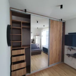 Duża szafa przesuwna z lustrzanymi drzwiami i drewnianymi panelami, z otwartą częścią z półkami i szufladami, odbijająca wnętrze pokoju dziennego z sofą i telewizorem.