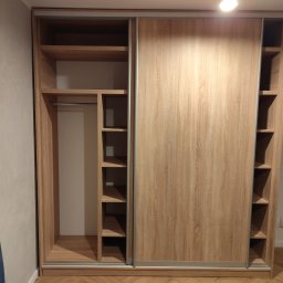 Michał Przybyszewski - Jasna szafa wnękowa z przesuwnymi drzwiami i półkami bocznymi, wykonana z płyty drewnopodobnej o naturalnym wzorze słojów.